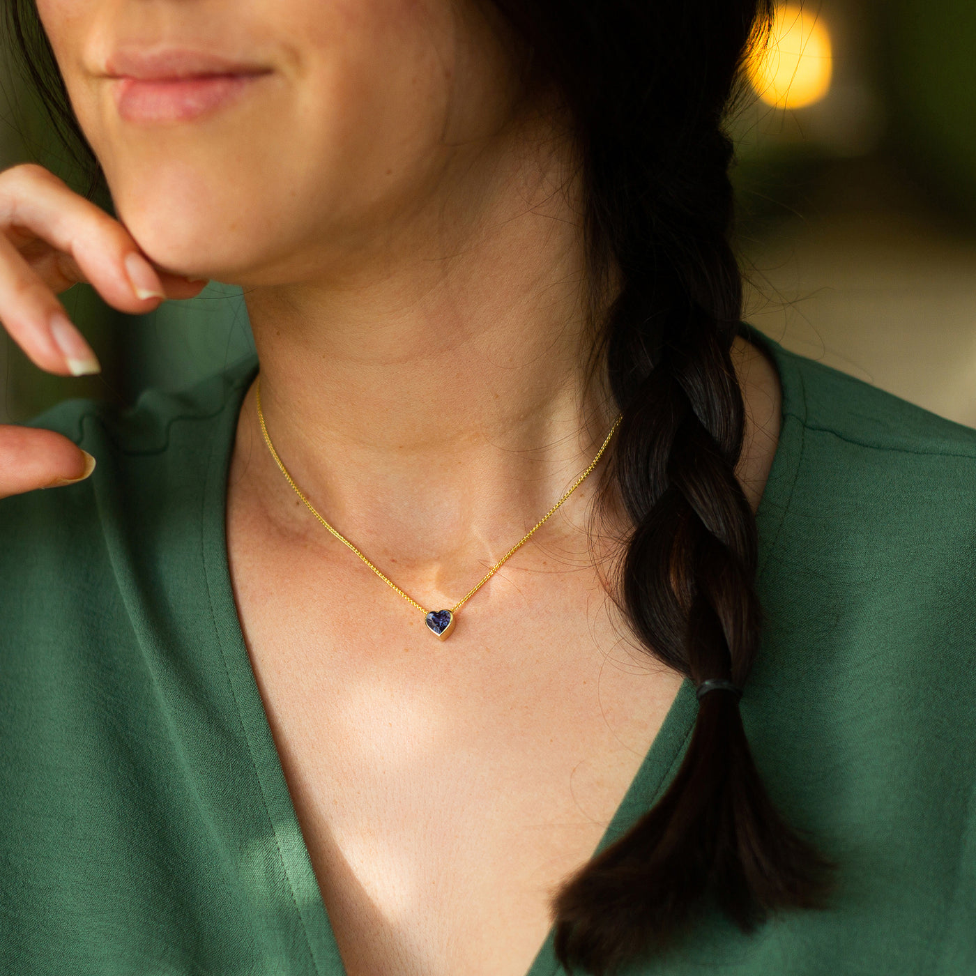 Woman wearing a gold necklace with a heart-shaped Alexandrite pendant, blurred background