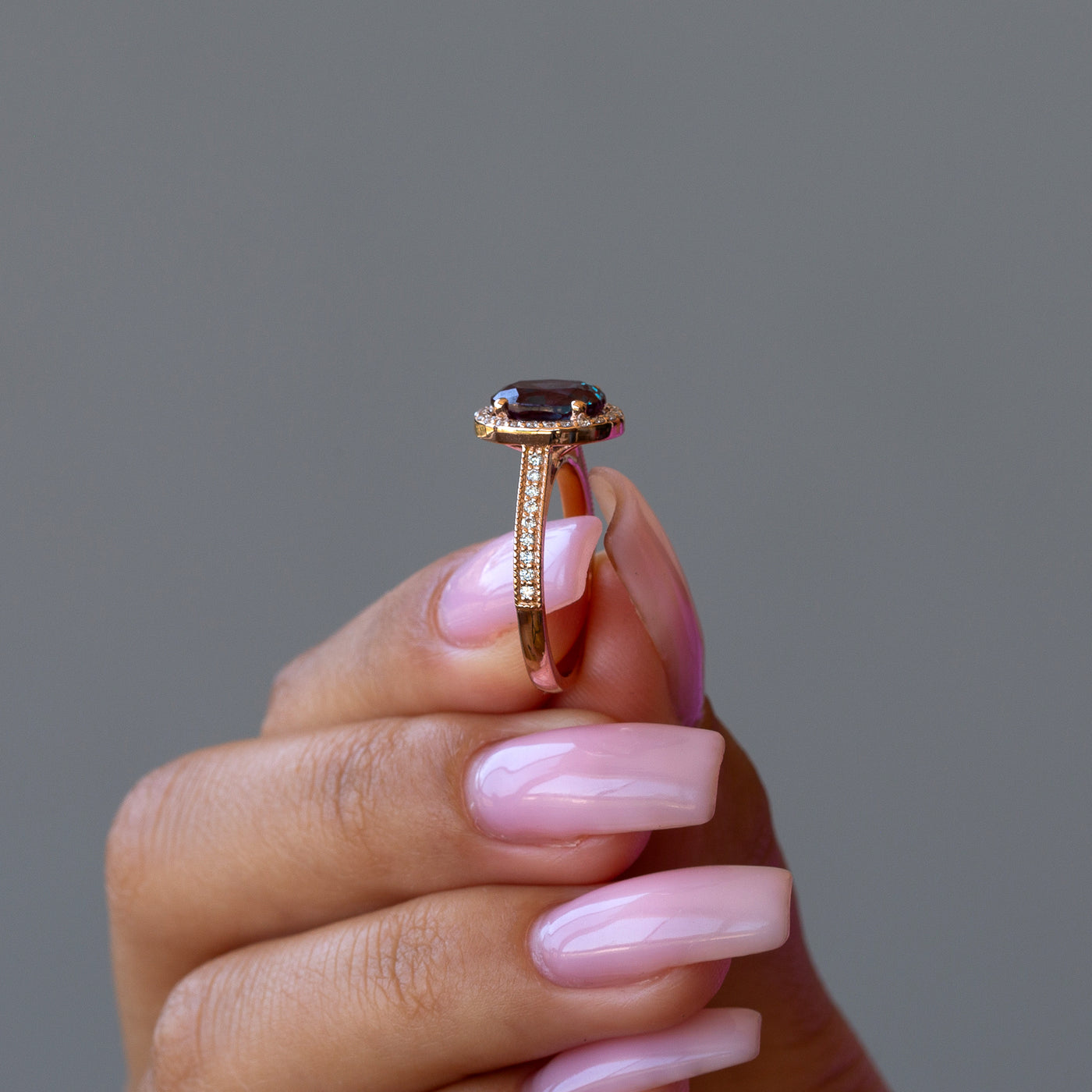 Gold ring with a teal gemstone held by a hand with pink nail polish against a gray background