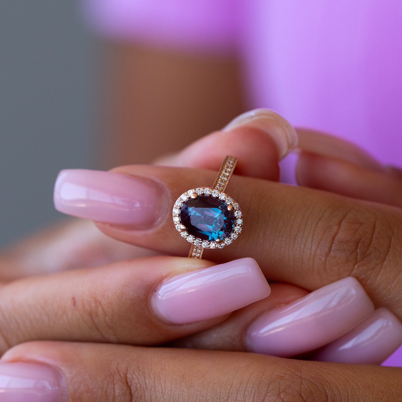 Close-up of a hand wearing an alexandrite engagement ring against a blurred background